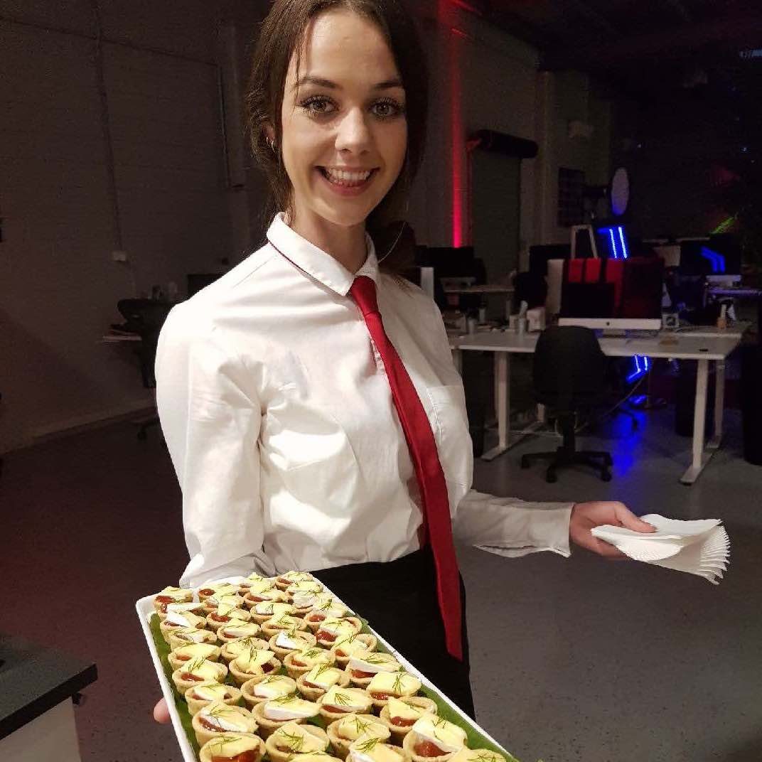 Smiling waitress dressed in white long-sleeved shirt with red tie holding platter of tartlets