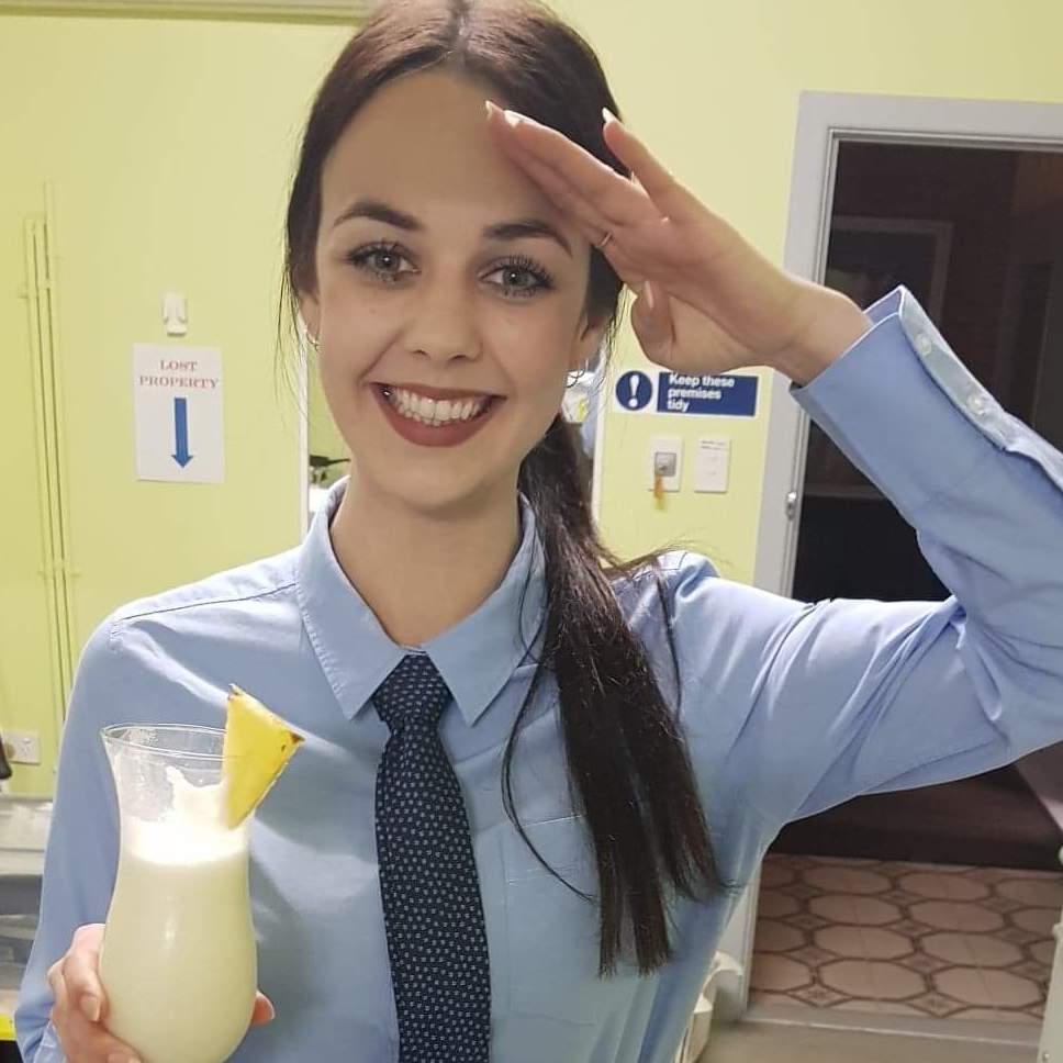 Smiling waitress dressed in light blue long sleeve shirt and tie saluting with a pina colada