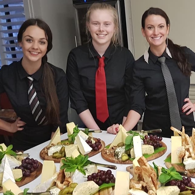 Three smiling waitresses dressed in black shirts and ties with six big cheese platters