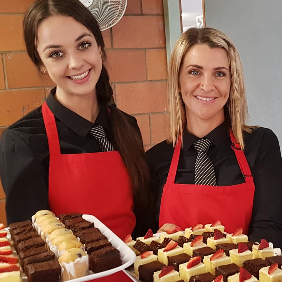 Two smiling waitresses dressed in black with red aprons holding dessert platters